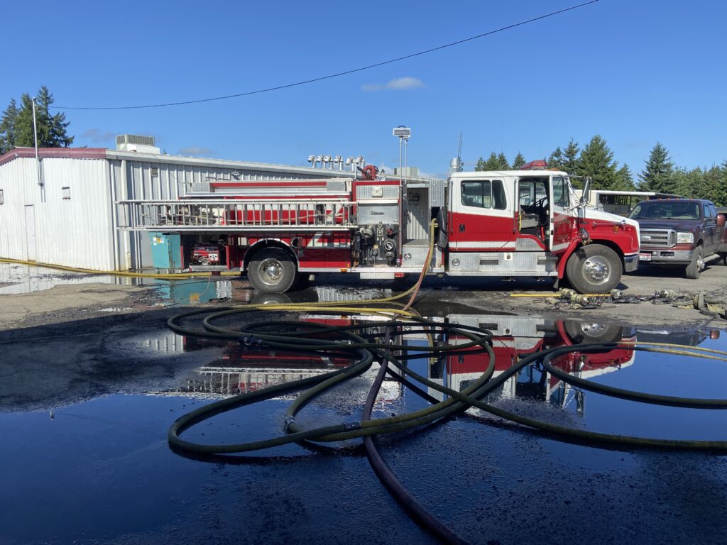 A red and white fire truck is positioned at an emergency scene with multiple large black hoses deployed across wet pavement. The fire truck appears to be a pumper truck with equipment compartments visible. Water puddles reflect the truck and clear blue sky above. In the background, there are industrial or commercial buildings with metal siding, evergreen trees, and power lines. Additional emergency vehicles are visible in the background. The scene suggests an active firefighting or emergency response operation.